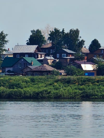 The image shows a residential area with several wooden houses situated along a riverbank in Yeniseysk, Russia. The houses have pitched roofs and appear to be of varying sizes and architectural styles, some with visible chimneys. The river in the foreground is calm with a slightly rippled surface, reflecting the light. The riverbank is covered in dense green foliage and trees. The sky is clear and pale blue. There are no people visible in the image.