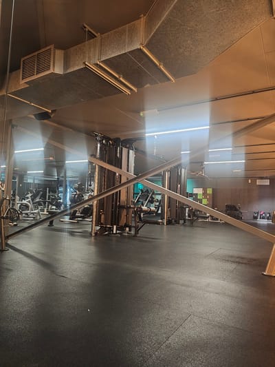 The image captures an indoor shot of a fitness center, likely located in Bristol, United Kingdom. The perspective is from an elevated position, looking down upon rows of gym equipment aligned on a dark, rubberized floor. Exercise machines dominate the scene, with various weightlifting and training stations visible.
In the background, there is a wall with light-colored lockers and a window. Above, there are ceiling-mounted fixtures and ductwork. The lighting in the room is a mix of fluorescent and natural light coming through the windows.
No people are discernible in the image. The overall impression is of a well-maintained and equipped fitness facility.