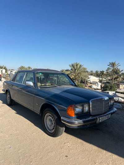 A vintage, dark blue Mercedes-Benz W123 sedan is parked on a sandy surface in Khan Yunis. The car is positioned at an angle, facing slightly to the right. The front of the car, including the grille, headlights, and chrome bumper, is visible. The license plate reads "3-0617-04". Behind the car, a cluster of palm trees and various tents and structures are visible, suggesting an outdoor or possibly temporary settlement environment. The sky is clear and blue. The ground is sandy with tire tracks and footprints visible.
