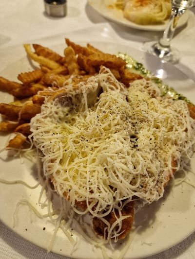 The scene displays a prepared meal on a white circular plate, situated on a white tablecloth in Odorheiu Secuiesc, Romania. The primary subject is a golden-brown, breaded cutlet, extensively covered with shredded white cheese and liberally dusted with ground black pepper. Adjacent to and partially behind the cutlet is a sizable portion of golden-brown, crinkle-cut french fries. A thin, wavy line of green material is visible along the right edge of the plate. The background, out of focus, reveals a metallic object (possibly a shaker) in the upper left quadrant, and the stem of a clear glass along with a section of a second white oval plate containing an indistinct light-colored item in the upper right. No individuals are present in the frame.