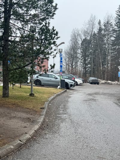 The scene depicts an outdoor parking area in Espoo, Finland, under an overcast sky. The ground is asphalt, partially wet or speckled with grit, and a concrete curb separates it from a grassy verge exhibiting brown and sparse green patches. Several vehicles are parked in parallel.

In the foreground, a light grey 4-door coupe/sedan, identifiable as a BMW i4, is parked on the left, actively charging via a white cable connected to a grey charging station on its front driver's side fender. To its immediate right, a dark grey hatchback is parked. Further right, a white sedan/hatchback is visible, followed by another dark vehicle partially obscured. In the far background, towards the right, a dark grey SUV is parked near a patch of residual snow.

A prominent blue parking sign with a white 'P' symbol is mounted on a light pole between the first two parked vehicles. Below the 'P', symbols indicate electric vehicle charging, disabled parking, and a right-pointing arrow. Multiple modern streetlights with flat-panel fixtures are visible throughout the parking area.

Vegetation includes mature evergreen pine trees in the foreground and forming a dense backdrop on the right, interspersed with bare deciduous trees. A multi-story brick-colored building is partially visible through the trees in the distant background on the left. No individuals are present in the frame.