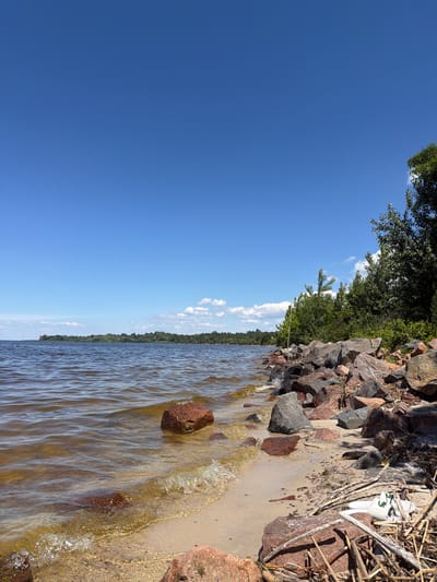 This is a photograph of a lake shore on a clear, sunny day. The sky is a vibrant blue with a few scattered white clouds. The lake water is a murky brown and appears calm with small ripples. The shoreline is composed of sand and a variety of large rocks and stones, some of which are partially submerged in the water. Dense green trees and foliage line the distant shore. In the foreground, a white plastic object, possibly a container or dish, is partially buried in sand and debris along the rocky edge. The scene is devoid of any human presence or activity. The location is identified as Lebedivka, Ukraine.