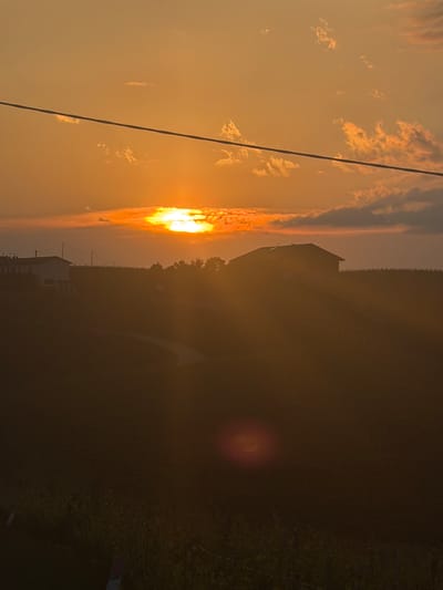 Captured at twilight, the image presents a partially obscured landscape with the sun positioned on the left, emanating an orange glow diffused by the atmosphere. A power line cuts diagonally across the sky. Silhouetted against the setting sun are buildings and trees, suggesting a rural or suburban setting. A road is vaguely visible. The location, specified as Castiglione Tinella, Italy, is not readily evident without further contextual information or visual landmarks within the image. No people are visible. The lighting is dim, suggesting either dawn or dusk, with a lens flare present.