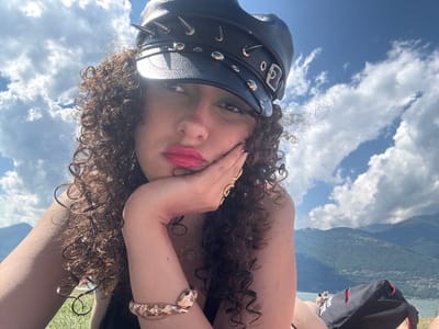 A woman with curly brown hair, wearing a black spiked hat and vibrant pink lipstick, rests her chin on her hand. She is outdoors, with a bright blue sky filled with fluffy white clouds above her. In the background, lush green mountains and a body of water are visible, suggesting a scenic location. A black bag with red trim is placed near her. In the distance, a few people can be seen near the water. The woman appears to be posing for a selfie, her gaze directed towards the camera. The overall impression is one of a relaxed, sunny day in a beautiful natural setting. The location is likely near the lake in Colico, Italy, given the mountainous backdrop and body of water.