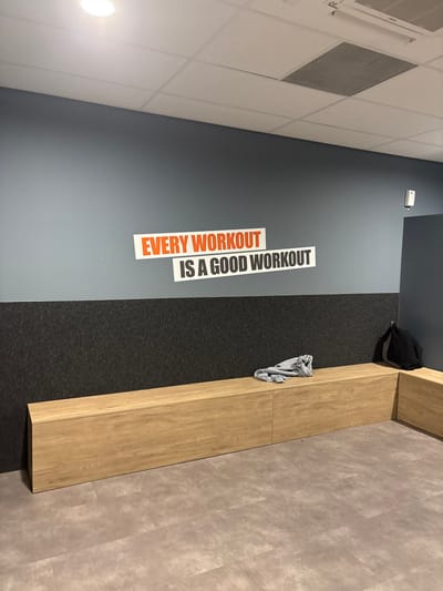 The image depicts an interior scene within a facility located in Rosmalen, Netherlands, likely a fitness center changing room or common area. The room features light grey-blue walls with a dark grey, textured paneling on the lower section. Above this paneling, a motivational quote is affixed to the wall, reading "EVERY WORKOUT" in orange text on a white background, positioned above "IS A GOOD WORKOUT" in black text on a white background. The ceiling is a white tiled false ceiling with a recessed circular light fixture on the left and a rectangular air vent/grille on the right. A light wood-toned, L-shaped bench unit extends along the wall on the floor. A crumpled light grey fabric item rests on the main section of the bench, and a black backpack is situated on the shorter, right-most section of the bench. The floor is covered with light grey-beige patterned vinyl or linoleum. A small white electronic device is mounted on the upper wall section near the right corner. No individuals are present in the scene.