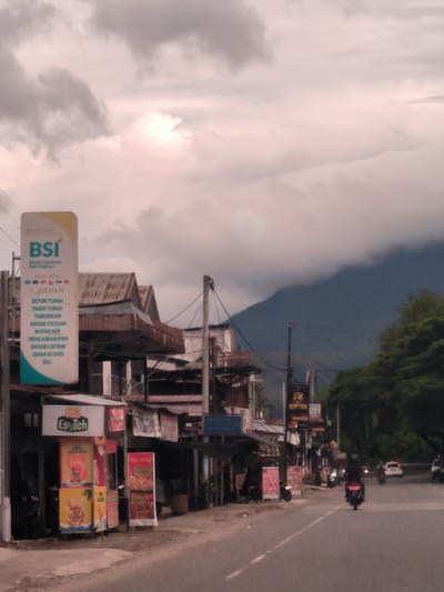 The image depicts a rural or semi-urban street scene in Reuhattuha, Indonesia, captured likely during a cloudy late afternoon or early evening. The road, a two-lane asphalt surface with a white center line, stretches into the distance, leading towards a large, misty mountain range in the background.
On the left side of the road, a series of simple, multi-story buildings with visibly rusty corrugated metal roofs house various small businesses. A tall, rectangular sign for "BSI BANK SYARIAH INDONESIA" stands prominently, advertising "MINI ATM LAYANAN" (Mini ATM services) including "SETOR TUNAI" (cash deposit), "TARIK TUNAI" (cash withdrawal), "TABUNGAN" (savings), "BAYAR CICILAN" (pay installments), "BAYAR SPP" (pay school fees), "PENCAIRAN PKH" (PKH disbursement), "BAYAR LISTRIK" (pay electricity), and digital payments via "DANA & OVO, DLL." Below, a stall advertises "Es Teh Poci" (iced tea) with "100% GULA ASLI" (100% real sugar). Further down, signs for "TIGA PUTRI," "Kentang Goreng" (French Fries), and "Jamur Crispy" (Crispy Mushrooms) are visible, along with a "RAPTOR" sign.
Activity on the road includes a person riding a black motorcycle in the foreground, moving away from the viewer. Other motorcycles and a white car can be seen further down the street. Overhead, numerous power lines and cables crisscross. The sky is predominantly overcast with thick, light grey clouds, and patches of brighter light suggest the sun is diffused behind them. Lush green trees line the right side of the road, partially obscuring the base of the dark, imposing mountain. The overall scene conveys a sense of everyday life in a mountainous region.