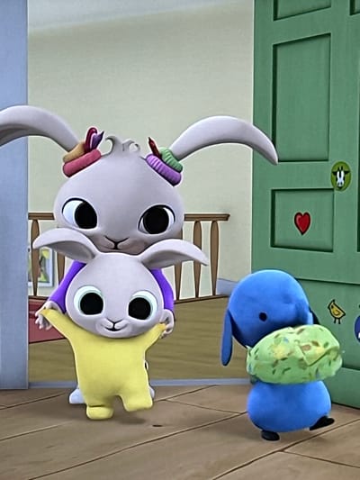 The image displays an animated indoor scene featuring three cartoon characters. A large light-grey rabbit, wearing a purple top and multicolored hair ties on its ears, stands on the left, supporting a smaller light-grey rabbit from behind. The smaller rabbit is dressed in a yellow full-body suit, has arms outstretched, and a slight smile. To the right, a small blue, elephant-like creature stands, holding a mottled green and yellow lumpy object with both hands. The background features a light-colored wall, a wooden railing with spindles, and a paneled green door. The green door is adorned with a round yellow-green sticker depicting an animal head outline, a red heart sticker, and two small bird-shaped stickers. The characters are positioned on a light brown wooden floor. The location of this depicted scene is Easingwold, United Kingdom.