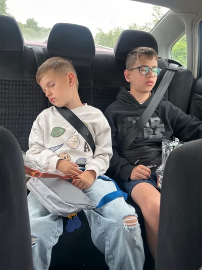Two young boys are asleep in the back seat of a car. The boy on the left is wearing a white sweatshirt with patches and light-wash ripped jeans. He is leaning his head against the headrest, with his eyes closed and mouth slightly open. A gray bag with blue straps is on his lap. The boy on the right is wearing a black Star Wars hoodie and blue patterned shorts. He has on glasses and is also asleep, leaning his head slightly forward. Both boys are wearing seatbelts. The car interior is visible, with the back of the seats and headrests. Outside the car windows, trees and greenery are visible, suggesting an outdoor setting, possibly a road in or near Benkovski, Bulgaria, given the context. The lighting suggests it is daytime, possibly overcast.