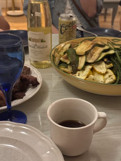 The image depicts a dining table scene, likely in Truro, Massachusetts, given the context of typical vacation spots in that region. In the foreground, a white ceramic mug holds dark liquid, possibly coffee. To the left, a blue-tinted glass, partially filled with a clear, bubbly beverage, stands on a pale blue plate. Adjacent to the glass, a white plate displays portions of what appears to be grilled steak.

Further back, a bottle of Chateau Ste. Michelle Dry Riesling wine, with a price tag of $12.99 visible, is positioned next to a can of Coors Light beer. A large, shallow bowl filled with grilled zucchini and asparagus slices sits prominently in the midground. The table surface is a light-colored, textured material, possibly wood or laminate, with a slight sheen. In the background, out of focus, a person is seated, wearing a denim jacket. The overall impression is of a casual meal shared among individuals, likely during a summer gathering.