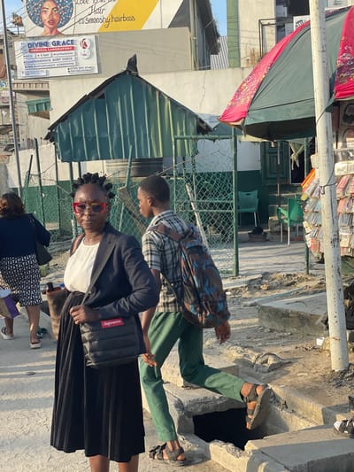 The image captures a street scene in Lagos, Nigeria, likely in a bustling commercial area. In the foreground, a woman with distinctive braided hair and colorful glasses is the central focus. She wears a dark blazer over a white top and a long, pleated black skirt. A black crossbody bag with a red "Supreme" logo is slung across her body. To her right, a young person wearing a plaid shirt and green trousers walks away from the camera, carrying a patterned backpack. The background reveals a mix of buildings and makeshift structures, including a green metal roofed building and a stall with various items displayed. A large pink and green umbrella is also visible, suggesting outdoor commerce or shelter. The ground is paved with concrete, and there are signs of urban disrepair, with a visible drainage opening in the foreground. The lighting suggests it is daytime, with sunlight casting shadows.
