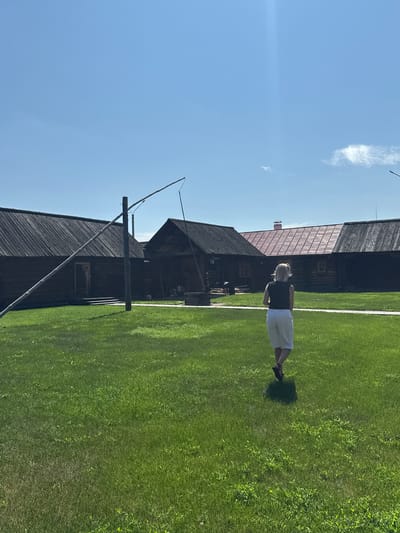 A woman with blonde hair, wearing a black sleeveless top and white shorts, walks away from the camera across a green grassy field. Behind her, a row of dark wooden, rustic buildings with sloped roofs stretches across the background. One building has a red-tiled roof. In the foreground, a wooden well with a long beam is visible, angled towards the left side of the frame. The sky is a clear, bright blue with a few wispy white clouds. The scene appears to be set in an outdoor historical or museum setting, possibly depicting a village in Shushenskoye, Russia.