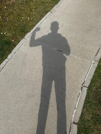 A single human shadow is cast upon a light grey concrete pathway in Radovljica, Slovenia. The shadow depicts a standing figure with its right arm bent at the elbow, hand clenched, suggestive of a bicep flex. The left arm is positioned alongside the body, with the hand visible near the hip. The concrete pathway exhibits visible expansion joints or cracks. To the immediate left of the pathway, a strip of green grass with scattered small white flowers and dried brown leaves is visible. A narrower strip of grass, bordered by a concrete curb, runs along the right edge of the pathway. The distinct shadow indicates strong overhead illumination from an unseen light source.