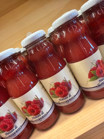 A close-up, angled shot displays several glass bottles of raspberry nectar. The bottles are filled with a vibrant red liquid and topped with white screw-on caps. Each bottle has a label featuring an illustration of raspberries and text that reads "Nettare di Lampone." The bottles are arranged on a light-colored wooden surface, likely a shelf or counter. The lighting is bright, creating reflections on the glass. The scene is indoors, possibly in a shop or pantry, in the city of Lateis/Latais, Italy. There are no people or animals visible in the image.
