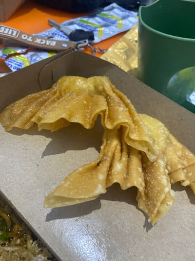 Two golden-fried wontons are presented in a cardboard takeout container. The wontons are arranged diagonally across the frame, with their pleated edges and slightly irregular shapes creating visual interest. The lighting casts a soft shadow to the left of the wontons. In the background, an orange surface is visible, with a keychain that reads "BACK TO LIFE" and a key attached, suggesting a casual setting. A green plastic cup and some foil packaging are also present. The foreground shows a glimpse of noodles, indicating this is a meal likely prepared or purchased in Walennae, Indonesia.