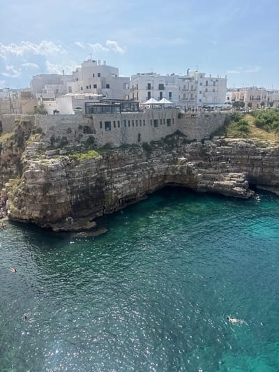 Here is a detailed description of the image:
The image shows a scene in Polignano a Mare, Italy, featuring a rocky coastline and turquoise sea. The camera angle is elevated, offering a view down towards the water. The water is textured with gentle ripples, indicating calm conditions, with several people swimming and sunbathing.
Adjacent to the water is a rugged cliff face, with exposed rock layers and patches of vegetation. Some people are sitting on the cliff.
On the left side of the image are closely built white buildings, a characteristic feature of Polignano a Mare. Some of the buildings appear to be built directly onto the cliffs, merging the man-made structures with the natural landscape. The sky is light blue with a few white clouds.