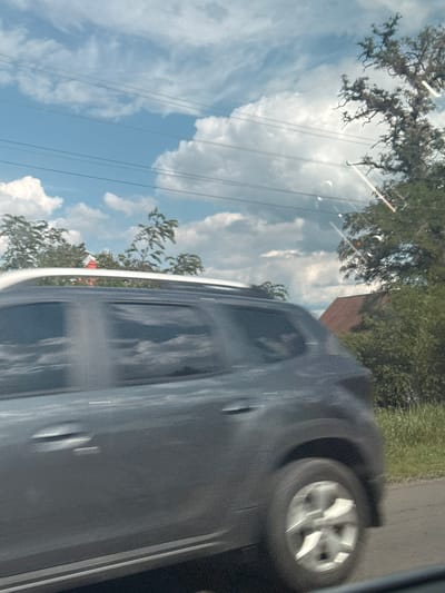 The image depicts a scene observed from a moving vehicle, likely in Markivka, Ukraine, during daylight hours. A dark grey mid-size SUV, equipped with white roof rails, is the primary subject, shown in motion from its passenger side and rear quarter, traveling from left to right. Significant motion blur is evident on the vehicle's body and its multi-spoke alloy wheels, which appear as blurred circular shapes. Reflections of the sky and clouds are visible on the SUV's darkened windows.

The background consists of a bright blue sky with scattered white clouds, including cumulus formations and wispy cirrus clouds. Multiple dark utility lines, appearing as five parallel horizontal strands, traverse the sky in the upper and middle portions of the frame. Various trees are visible along the roadside; on the left, blurred green foliage is present, while on the right, a large tree with pronounced, gnarled bare branches stands against the sky. Further right and partially obscured by trees, a building with a distinct red, gabled roof is discernible. The immediate foreground on the right side of the road shows dark asphalt and green roadside vegetation. Several bright, streak-like lens flares and circular reflections are present, particularly in the upper right quadrant of the image, consistent with light interacting with a car window or camera lens. No specific individuals are clearly identifiable within the frame.
