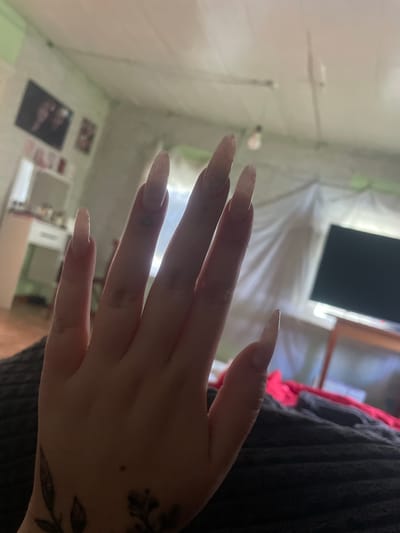A close-up, low-angle shot shows a left hand with long, light-colored, pointed acrylic nails extending towards the viewer. The hand is positioned in the foreground, obscuring the lower portion of the image. In the background, a room is visible with a television on the right, a blurred dresser with items on it to the left, and a light green wall. A light fixture with a bare bulb hangs from the ceiling. The scene appears to be an indoor domestic setting in the city of Linovka, Russia.