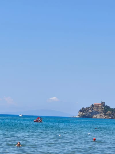 A clear, bright summer day unfolds over the waters near Castiglione della Pescaia, Italy. The azure sky is vast and cloudless, with a few wispy formations in the distance. The deep blue sea, calm and inviting, stretches towards the horizon.
In the foreground, a young person with dark hair is partially submerged in the water, facing forward with a slight smile, wearing headphones. Further out, a small red paddleboat is occupied by at least two individuals, one steering and another standing. A white motorboat is visible in the middle ground.
Dominating the background is a rocky promontory topped by a fortress-like structure with red-tiled roofs. This iconic landmark is situated on the coast, with the rugged cliffs dropping sharply to the sea. The distant coastline is a faint, hazy line of mountains.
Several buoys mark an area of the water, including a bright orange one and two white ones. The overall scene depicts a tranquil day at the beach, with recreational activities taking place in the sea against a backdrop of historical architecture and natural beauty.