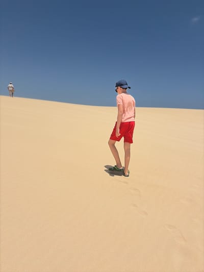 This vibrant image depicts a vast, light-colored sand dune stretching under a clear, brilliant blue sky in La Oliva, Spain. The horizon line diagonally bisects the frame, separating the expansive sandy terrain from the deep azure sky.

In the mid-ground, a young person, possibly a boy, stands prominently on the dune. He is dressed in a light pink t-shirt, red shorts, a dark blue baseball cap, and green and black sandals. His head is slightly tilted downwards, and he appears to be looking towards his left (when viewed upright). The soft sand around him shows numerous footprints, suggesting recent activity or exploration.

Far in the distance, close to the horizon on the left side of the frame, a smaller, indistinct figure dressed in light-colored clothing is also visible, standing on the dune. The overall scene is brightly lit by natural sunlight, indicating a clear and sunny day. There is no visible text in the image.