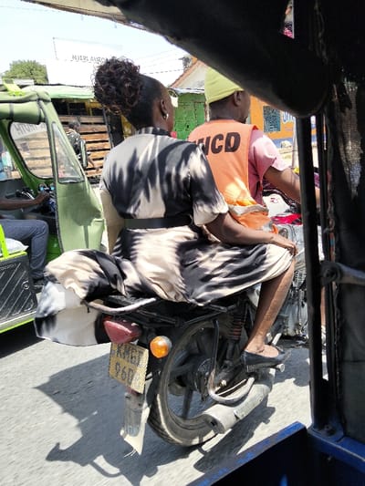 An outdoor street scene in Mkomani ward, Kenya, depicts an adult female passenger riding sidesaddle on the rear of a motorcycle, driven by an adult male. The woman, viewed from behind, has dark hair styled in a bun and wears a black and beige/white patterned short-sleeved dress with a black belt, and black flat shoes. The male driver, also viewed from behind, wears a yellow cap, a pink short-sleeved shirt, and an orange high-visibility vest with "NCD" visible on the back.
The motorcycle is dark-colored, featuring a visible rear wheel, exhaust pipe, suspension, a red taillight, and an amber turn signal. A yellow Kenyan license plate, partially obscured but showing "KMG" and "96D", is attached to the rear. To the left of the motorcycle is a green tuk-tuk (auto-rickshaw), with its driver partially visible inside. The image is framed from within another vehicle, likely a dark-colored tuk-tuk, evident by the dark structure in the foreground.
The environment is an urban setting under bright daylight, indicated by sharp shadows on the paved road. Background elements include various single-story buildings with corrugated metal roofs, some painted in bright colors like green and blue. A white sign with "NJERI AUTO" is visible above the green tuk-tuk. Both the motorcycle and the green tuk-tuk appear to be either in motion or paused in traffic.