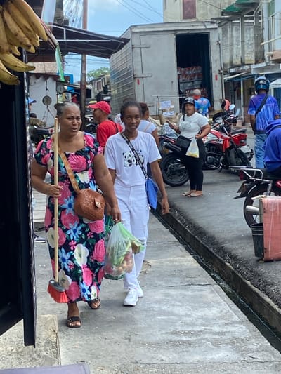 In San José de Barlovento, Venezuela, a street scene unfolds with several individuals engaged in everyday activities. The foreground features two women walking on a concrete sidewalk. The woman on the left, wearing a floral dress, carries a broom and a brown crossbody bag, while the woman on the right, dressed in white, holds a clear plastic bag filled with produce and wears white sneakers. Behind them, a red motorcycle is parked. Further back, a large truck with its cargo doors open reveals stacked boxes, suggesting a delivery or distribution point. Other people are visible in the background, including a man in a red cap and another wearing a blue shirt with a lightning bolt pattern. The environment appears to be an outdoor market or commercial street, characterized by buildings, overhead structures, and a general atmosphere of bustling activity. A cluster of bananas hangs from a structure on the far left.