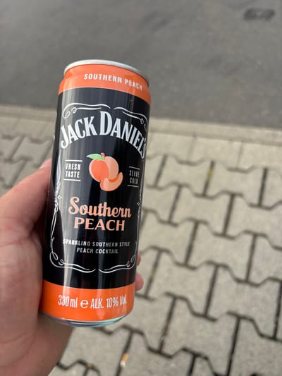 A person holds a black and orange can of Jack Daniel's Southern Peach sparkling cocktail. The can is oriented vertically, with the top facing upwards. The label features the Jack Daniel's logo in white, with "Southern Peach" written in a stylized font below an illustration of two peaches. Text on the can indicates "Sparkling Southern Style Peach Cocktail," "Serve Cold," "Fresh Taste," and "330 ml e ALK. 10% Vol." The background shows a textured grey surface, possibly a paved walkway with interlocking concrete stones. A discarded cigarette butt is visible in the upper right corner of the frame. The lighting suggests it is daytime, outdoors. The image appears to be taken in Elsenfeld, Germany.