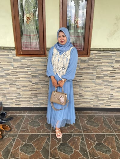 A single adult female stands centrally on a tiled patio or porch in Ketegan, Indonesia. She wears a light blue hijab, a long-sleeved light blue dress, and a cream-colored crocheted vest. Her footwear consists of light pink, open-toed, multi-strap sandals. She holds a light brown handbag, featuring a gold chain detail and a patterned scarf, with both hands at waist level. A gold-toned watch is visible on her left wrist. The background consists of a wall, with the lower half constructed from horizontal, stacked stone veneer in shades of grey and beige, and the upper half a plain light cream-colored painted section. Two dark wooden-framed windows with patterned translucent curtains are visible in the upper background, reflecting external greenery. The floor is covered with irregular, polygonal brown and dark grey tiles. Two pairs of casual sandals, one black and one brown, are visible on the floor to the left of the subject.