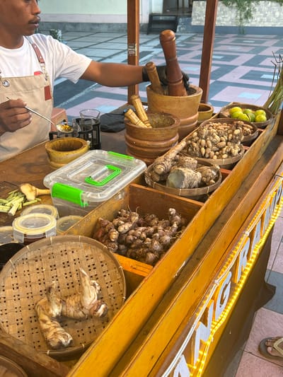 A person, identified as a male wearing a white t-shirt and a tan apron, is preparing ingredients at a wooden food stall located in Bantaeng, Indonesia. The stall is filled with various spices and herbs in woven baskets, including what appears to be galangal, turmeric, ginger, and lime. There are also wooden mortars and pestles, suggesting the preparation of traditional Indonesian herbal remedies or spices. The stall has a wooden frame and is decorated with bright yellow LED lights that spell out words. The background shows a checkered tiled floor and some greenery, indicating an outdoor setting. The person is holding a spoon and a small glass, possibly in the process of extracting or mixing a liquid ingredient.