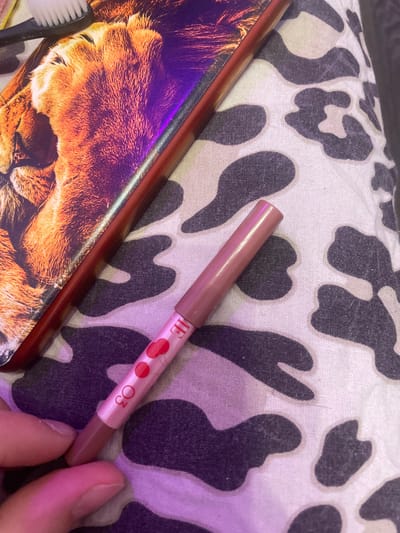 A hand is holding a light pink lip liner pencil with red heart designs and the numbers "03" visible on it. The pencil is positioned diagonally across a white fabric with a black and white cow print pattern. To the upper left of the pencil, partially visible, is the edge of a rectangular object with a close-up image of a lion's face and iridescent purple detailing. A white toothbrush is also visible in the upper left corner, resting on the lion-themed object. The scene appears to be indoors with artificial lighting casting a pinkish hue on some areas. There is no discernible information about the location within the city of Romanovka, Russia.