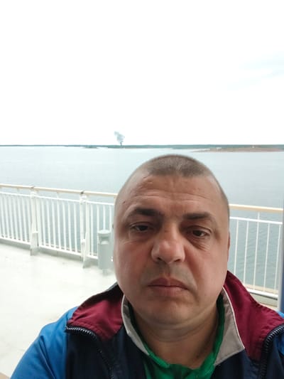 This is a selfie taken by a man on the deck of a ferry. He is wearing a blue and maroon jacket over a green shirt. The man's face fills the foreground of the image. Behind him, a white railing runs along the edge of the deck, separating it from the open water. The water is calm and gray, and the sky is overcast. In the distance, a smudge of smoke rises from the land on the horizon, suggesting industrial activity or perhaps a natural phenomenon. The overall impression is one of a cloudy day at sea, with the man capturing a moment on his journey. The setting appears to be Näset, Åland, a location known for its archipelago and ferry connections. There is no visible text in the image.