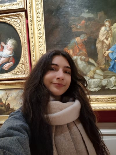 An individual with long, dark, wavy hair, approximately 18-25 years of age, is depicted in a half-profile selfie, smiling directly at the camera. She wears a grey denim jacket and a light beige knitted scarf. The background consists of a wall displaying multiple framed oil paintings, indicating an art museum or gallery. The frames are ornate and gold-colored, with sections of dark red wall visible between them. To the upper left, an oval-framed portrait shows a fair-skinned infant or young child with blonde, curly hair, held by an unseen figure. To the upper right, a larger rectangular painting portrays a multi-figure religious scene, consistent with a Lamentation or Deposition of Christ, featuring a pale, deceased male figure on a white sheet, surrounded by several draped individuals, including a kneeling male and a standing female. A smaller, partially visible landscape painting is also present in the lower-left background. This scene is located within Schloss Johannisburg (Johannisburg Palace), specifically the Staatsgalerie Aschaffenburg, in Aschaffenburg, Germany.