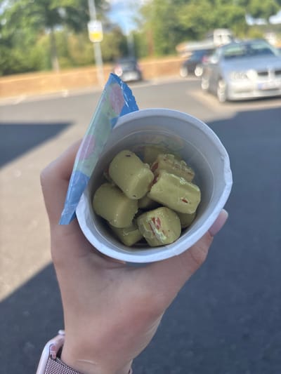 A person's hand holds a white paper cup filled with small, pale green, bite-sized candies. These candies appear to have a reddish interior visible in some of them, suggesting they might be filled or have a contrasting coating. The cup's lid, a blue and white plastic wrapper with colorful abstract designs, is partially open and hangs down the side.
The background is blurred, but it appears to be an outdoor setting, possibly a street or sidewalk. Sunlight casts a shadow on the ground, indicating it is daytime. In the distance, there are out-of-focus vehicles and greenery, suggesting an urban or suburban environment. The overall mood conveyed by the image is casual and perhaps indicates a moment of enjoyment or a snack break. There is no visible text on the candies or the cup itself. The crops highlight the candies and the cup's packaging.