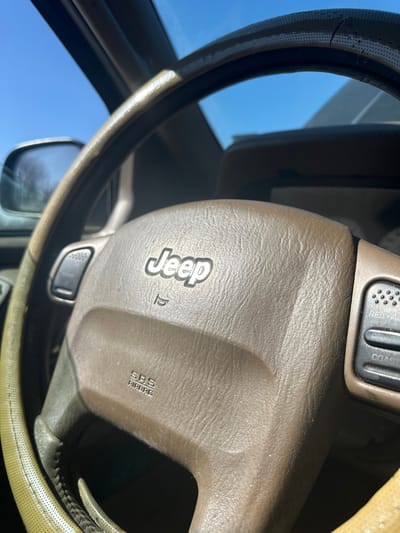 The scene depicts the interior of a vehicle, specified as being in the city of Golyanovtsi, Bulgaria. The primary subject is the brown steering wheel, which is partially covered by a light beige, worn, perforated cover. The central airbag module of the steering wheel features the "Jeep" brand logo prominently, with a small horn icon below it and "SRS AIRBAG" text embossed at the bottom. To the right of the steering wheel hub, a set of dark grey control buttons includes visible labels for "RESUME" and "COAST," indicative of cruise control functions. A partial view of a dark grey control panel is visible on the left side of the hub. Through the vehicle's windshield, a clear blue sky is visible, suggesting an outdoor, daylight environment. The left side mirror is partially visible, reflecting a blurred image consistent with distant trees or foliage. The vehicle's dashboard is visible in the background, appearing to be a consistent brown or tan color. No individuals are visible.