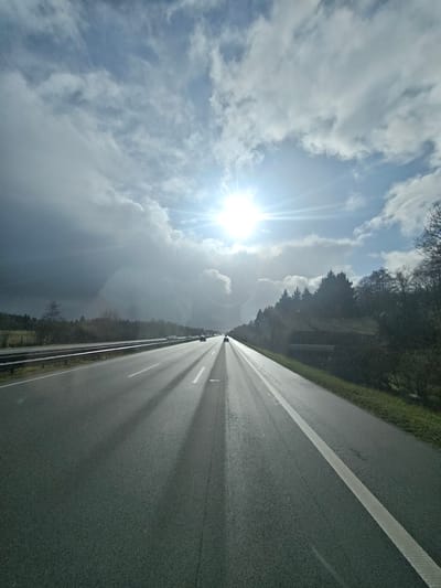 A medium-shot captures a sunlit highway somewhere near Heidenau, Germany. The road, flanked by trees and grass on the right, stretches towards the horizon, where a cluster of cars can be seen. The sky above is a mix of bright sun and looming, dark clouds, casting long shadows across the road. A metal guardrail runs alongside the highway's left side, adding to the scene's sense of location.