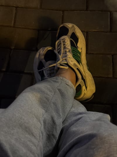 In an outdoor setting, likely a sidewalk or pedestrian area paved with dark, rectangular bricks, a person's legs are visible. The left leg is crossed over the right, with the right foot adorned in a white and green athletic shoe. The shoe features laces and a distinct design with green accents. The person is wearing grey sweatpants. The lighting suggests it is evening or night, with shadows cast on the brick surface. The specific location of Doma 1096 Km, Russia, is noted, though no visual cues in the image directly confirm this.