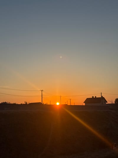 A silhouette of a residential building with a gabled roof and two chimneys is visible against an orange and blue gradient sky at sunset. The sun, a bright orb, is low on the horizon, casting a golden ray across a dark, grassy embankment in the foreground. Several utility poles with wires are scattered across the middle ground, with some trees and a fence line behind them. The scene appears to be in a rural or suburban setting.