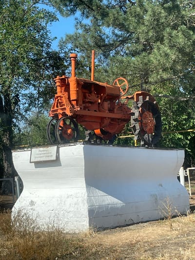 An outdoor scene shows an orange tractor on display next to a white monument. The tractor is old and appears to be made of metal, with large wheels. It is positioned at an angle, with its engine and front part more visible. The monument is a large, rectangular structure painted white, with some texture on its surface. A small plaque is attached to the monument, with some text visible. The scene is surrounded by greenery, including trees with pine needles and other foliage. The sky is visible through the trees and appears to be blue, suggesting a clear day. The overall impression is that of a monument or exhibit dedicated to agricultural machinery. The dry grass on the right side of the image may suggest a rural or natural setting. The text on the plaque appears to be in Cyrillic script.