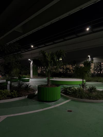 An outdoor space at night features green circular pathways lined with white. Tropical plants grow in raised planters, some cylindrical and green with horizontal slats. A large overpass looms overhead, its dark underside contrasting with the illuminated greenery. A streetlamp casts a glow on the scene, highlighting the blend of nature and infrastructure.