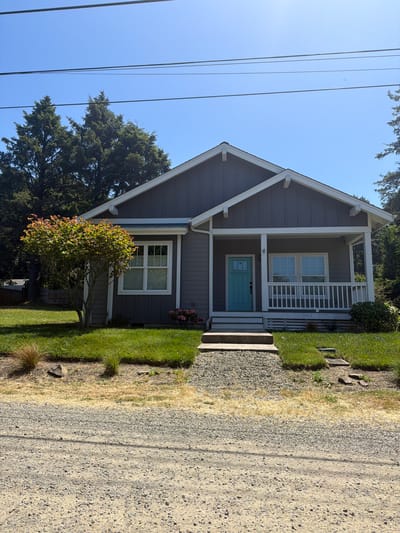 The image shows a residential house in Cannon Beach, United States of America, on a clear, sunny day. The house is a single-story structure with a grey exterior and white trim. It features a prominent front porch with white railings and a bright teal front door. There are two windows on the front of the house, one to the left of the porch and one to the right. A small tree with reddish-green leaves stands to the left of the porch, and a patch of pink flowers is visible near the base of the house.
The house is situated on a grassy lawn, with a gravel driveway leading up to it. The foreground of the image is dominated by the gravel driveway, which appears to be unpaved. In the background, tall evergreen trees form a dense backdrop. Power lines are visible stretching across the clear blue sky above the house and trees. The overall scene suggests a suburban or rural setting, characteristic of Cannon Beach. There are no people visible in the image.
