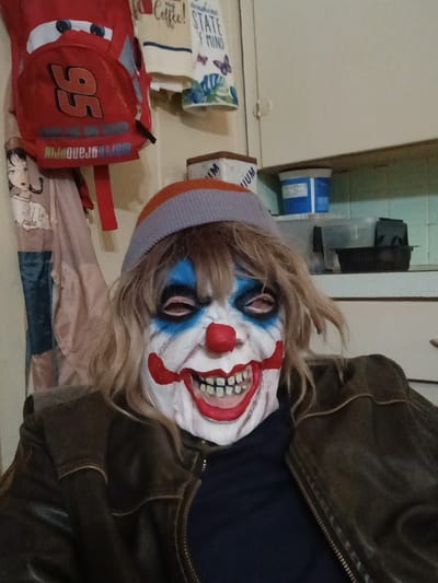 A person wearing a clown mask and a beanie is in a room. The mask is white with blue around the eyes, red lips and nose, and painted teeth. The person is wearing a brown jacket. Behind the person, there is a bed with a pillow and a red backpack with the number 95 on it. There is also a roll of paper towels and a blue container on a shelf. The location is in a room in a building in the city of Clarksville, United States.