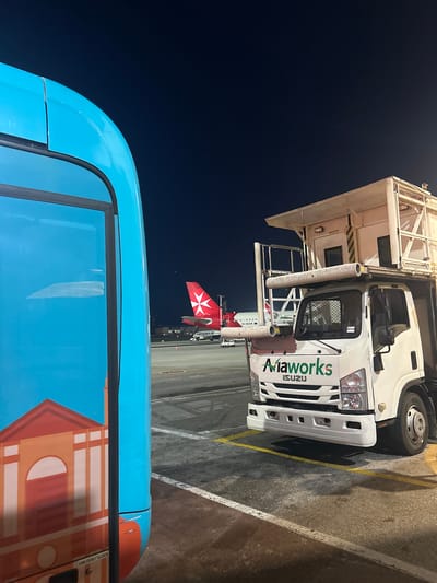The image depicts an airport scene in Luqa, Malta, at night. A white Isuzu "Aiaworks" catering truck with a platform is parked on the tarmac. In the background, a Ryanair aircraft with the Maltese eight-pointed cross on its tail fin is visible. The foreground features a blue bus with a graphic of a building. The ground is paved with asphalt, and there are painted yellow lines indicating parking. The sky is dark.