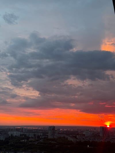 The image depicts a sunset over the city of Savenki, Russia. The sky is dominated by vibrant hues of orange and red, with dark clouds scattered throughout. The sun, a brilliant ball of light, is partially obscured by a building on the right side of the frame. Below the sky, a cityscape stretches across the horizon, featuring a mix of residential buildings and taller structures. Trees and greenery are visible in the foreground, suggesting a park or wooded area within the city. The overall scene is one of a dramatic and colorful end to the day.