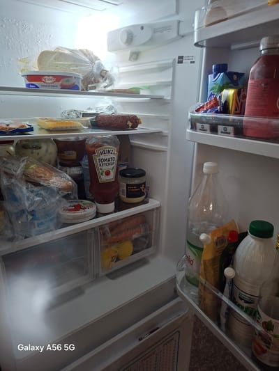 This media file shows an inside view of a refrigerator. The main subjects are various food and drink items, including bottles of ketchup, mayonnaise, juice, and other condiments, as well as packaged cheese, bread, and what appears to be salami. The scene is set inside a refrigerator, indicated by the shelves, drawers, and white interior walls. There is no activity or event taking place beyond the storage of food.
Notable details include the presence of several brands, such as Heinz, President, Kikkoman, and Sainsbury's. The text "Galaxy A56 5G" is also visible, suggesting that the photo was taken with a Samsung Galaxy A56 5G phone. The location context is Greater London, United Kingdom. There are no discernible cues about the time of day or weather. The overall impression is a well-stocked refrigerator.