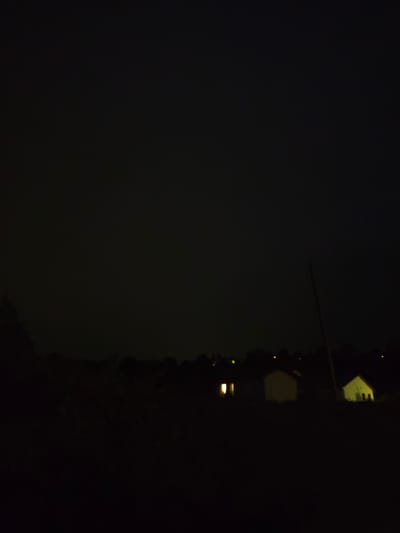 The image depicts a nighttime scene in Guarene, Italy. The sky is dark and overcast, with no visible stars or moon. In the foreground, silhouetted against the dark sky, are several small houses or structures. One structure on the left has two illuminated windows, suggesting interior lighting. Another structure in the center is unlit, appearing as a dark shape. To the right, a third structure has an illuminated doorway and window. A tall, thin pole, possibly a utility pole or tree, stands vertically on the right side of the frame, extending from the ground into the sky. The surrounding environment appears to be rural or on the outskirts of a town, with dark, indistinct shapes suggesting trees or vegetation. The overall impression is one of a quiet, nocturnal landscape.