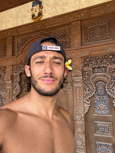 A man wearing a dark-colored Boston Red Sox baseball cap with a yellow flower behind his ear is seen in a selfie-style image. He has dark hair and a beard. The background features intricate wooden carvings, with a small, colorful figurine visible in the lower left corner. The carvings appear to be part of a door or wall. The man's chest is bare, and the overall lighting suggests an indoor setting.