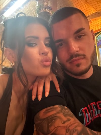 A forensic examination of the image reveals two individuals, a female and a male, captured in a close-up, selfie-style photograph within an indoor establishment in Debrashtitsa, Bulgaria.
The female, positioned on the left and closer to the camera, is an adult with dark hair, styled with strands framing her face. She wears full facial makeup, including defined eyebrows, dark eyeliner, and eyeshadow. Her lips are pursed in a "kiss face" expression, and her gaze is directed towards the camera. She is adorned with gold-toned hoop earrings, a thin gold-toned necklace featuring a bar pendant, and a thin gold-toned ring on her left ring finger. Her fingernails are long, oval-shaped, and appear to have a light-colored or clear polish. She is wearing a dark, possibly black, halter or tank-style top. Her right hand is visible, resting on the male's left shoulder/upper arm area.
The male, positioned to the right and slightly behind the female, is an adult with a short, buzzed dark haircut. He has a neatly trimmed goatee and mustache, with a short beard along his jawline. His expression is neutral, and his gaze is directed towards the camera. He is wearing a black t-shirt with partially visible embroidery on the right chest area, featuring red and blue lettering that appears to spell "DIES." His left arm is prominently displayed, covered by a detailed tattoo sleeve depicting a stylized face, possibly a woman with a bandana, among other patterns.
The background indicates an indoor setting with warm, artificial lighting. A long, bright light fixture is visible on a wooden-paneled ceiling at the top left. Decorative elements include a white, ornate, possibly wire-frame object with green foliage intertwined, visible at the top right. To the far right, a brightly lit, colorful screen or panel, possibly from an arcade machine or similar entertainment device, is partially visible. The overall environment suggests a commercial establishment such as a restaurant, bar, or entertainment venue.
