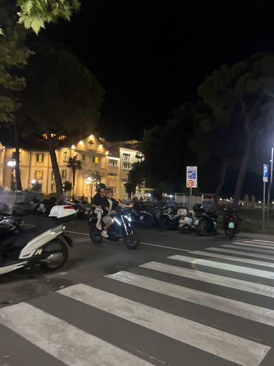 The image captures a nighttime street scene in Sestri Levante, Italy, focusing on a road segment with a marked pedestrian crosswalk. In the midground, two individuals are riding a dark motorcycle from right to left across the white-striped crosswalk. The driver wears a dark helmet and dark clothing. The passenger, positioned behind the driver, wears a light-colored top, light shorts, white sneakers, and sunglasses, with their right arm extended. To the immediate right of the motorcycle, a densely packed row of parked scooters and motorcycles occupies a designated area. A white rectangular sign displaying a red circle with "30" (indicating a speed limit) is visible above the parked vehicles, alongside other smaller signs. Further right, a blue square sign with a white "P" (parking) is present.
In the background, on the left, a multi-story building with illuminated windows and a distinctive peaked architectural feature is prominent, suggesting a hotel or large residential structure. Another building is partially visible to its right. Tall, mature trees and palm trees are interspersed throughout the scene, silhouetted against the dark night sky, with some foliage extending into the upper left foreground. Streetlights illuminate the road and surrounding areas. A white scooter is partially visible in the lower left foreground, near the crosswalk.