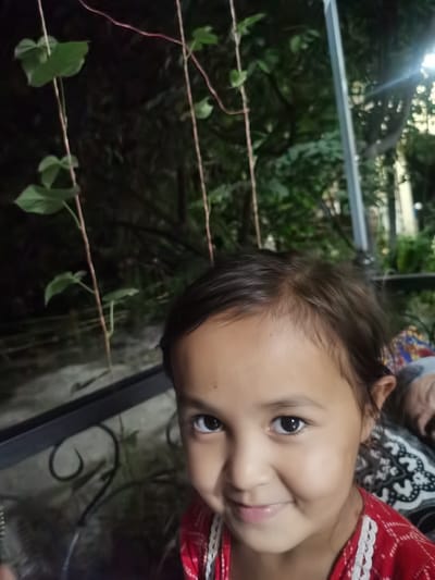 In Karabair, Uzbekistan, a young girl with dark hair and brown eyes smiles at the camera. She is wearing a red shirt with white trim. The background is dark and out of focus, but appears to be an outdoor setting with trees and greenery. A metal railing is visible in the foreground.