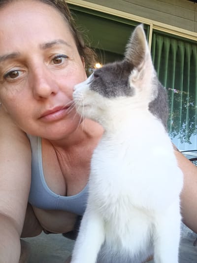 A woman poses for a selfie with a gray and white cat. The woman has brown hair pulled back and is wearing a gray tank top. The cat is facing to the right. Behind them are green curtains and an exterior wall. The photo was taken in Ban Tak Daet, Thailand.
