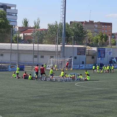 The image depicts an outdoor soccer field in Zaragoza, Spain, with several individuals, primarily children, engaged in a practice or training session. The field is artificial turf with visible white lines marking boundaries and a penalty area. In the background, there are buildings, trees, a tall metal structure resembling a stadium light or observation tower, and a chain-link fence surrounding the field.

There are at least 15 individuals visible, mostly wearing bright yellow or green t-shirts and dark shorts, suggesting they are part of a team or academy. A few adults are also present, wearing red t-shirts and dark shorts, likely coaches or supervisors. Some children are sitting on the ground, while others are standing or in motion. There are several portable white benches or bleachers arranged on the field, and a small portable soccer goal is visible on the right side.

One child in a yellow t-shirt is sitting on the ground with their legs extended, facing left. Another child in a similar outfit is standing next to them. Two adults in red shirts are standing further back, one facing the camera with hands on hips, and the other looking towards the soccer goal. Several children in yellow are gathered around the benches. One child in a red shirt appears to be in motion near the goal.

The environment is sunny with a clear blue sky. Advertisements are visible on the wall behind the fence, including one that reads "REPARALIZARA" and another with "MOTOR BOX". There is also a sign with text that includes "Pinturas de Aragón, S.L." and a phone number, indicating a local business presence. The overall scene suggests a youth soccer training event on a well-maintained sports facility.